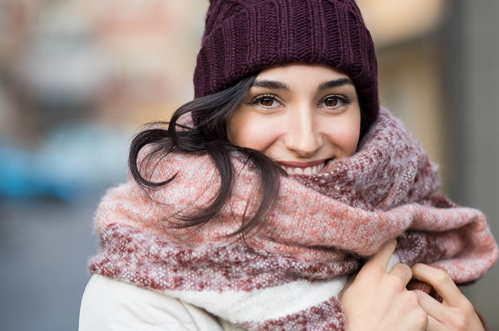 Closeup face of a young happy woman enjoying winter wearing scarf and cap. Smiling girl in a colorful shawl looking at camera. Latin woman with knitted bordeaux hat and woolen scarf.