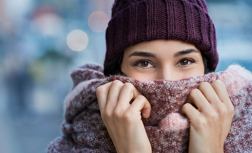 Winter portrait of young beautiful woman covering face with woolen scarf. Closeup of happy girl feeling cold outdoor in the city. Young woman holding scarf and looking at camera.