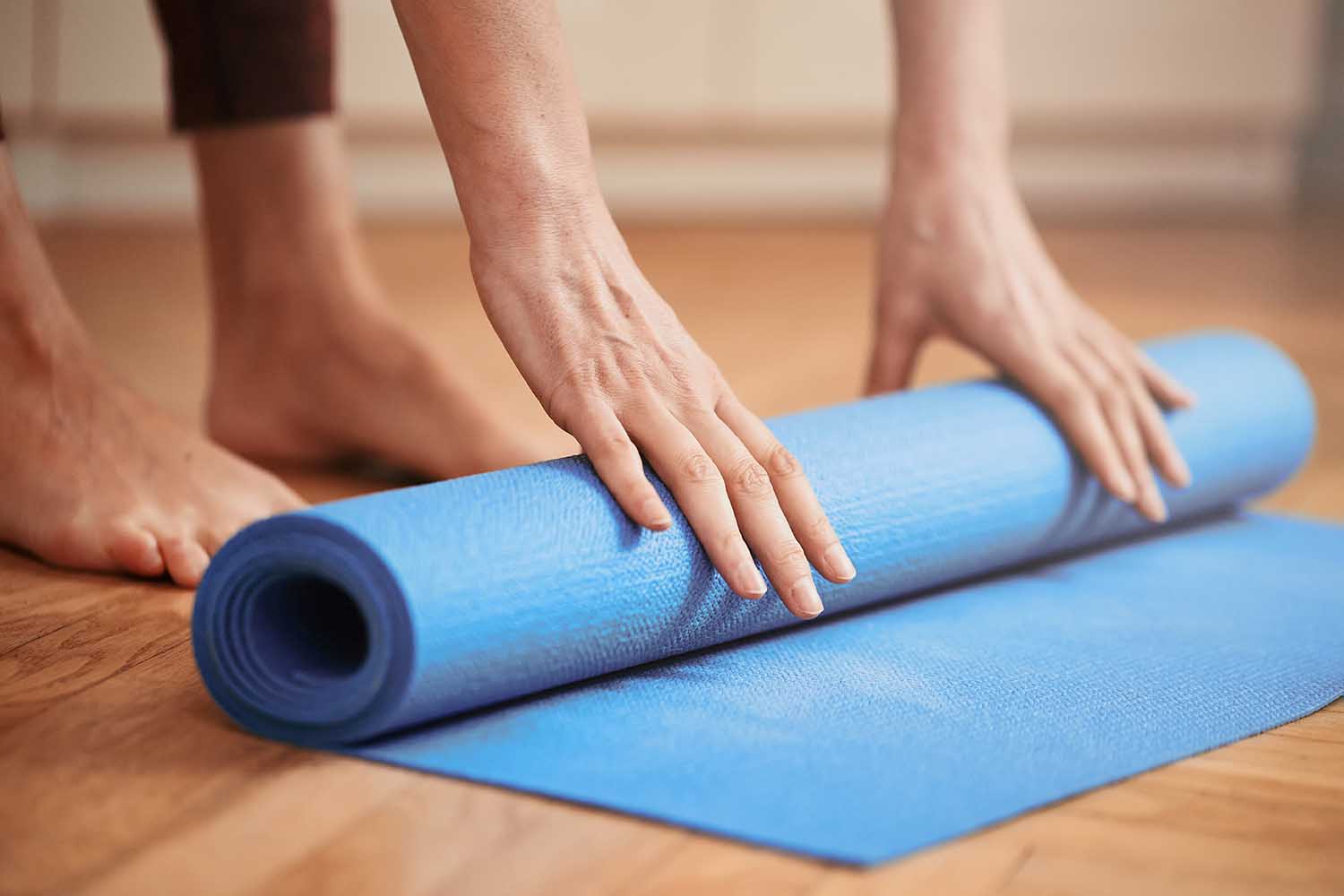 Close up of mature woman folding blue yoga or fitness mat before or after working out at home in living room. Healthy life in covid-19 time lockdown. Focus on hand. Little bit of film grain