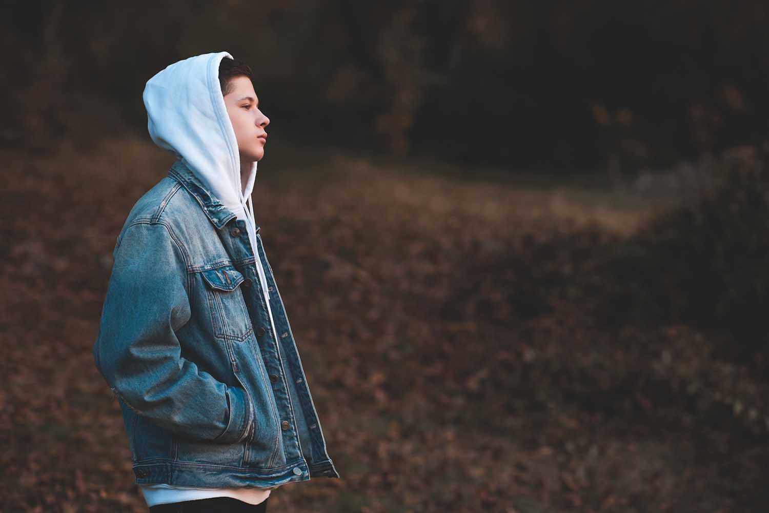 Handsome teen boy wearing denim jacket and white hoodie standing outdoors over nature autumn background. Teenagerhood.