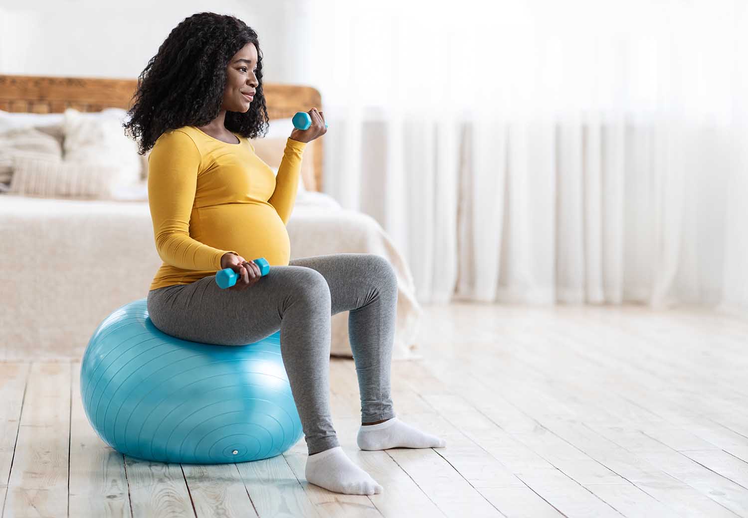 Active pregnant black lady exercising with barbells while sitting on fitness ball and looking at copy space, bedroom interior. African american expecting lady training at home, using yoga ball
