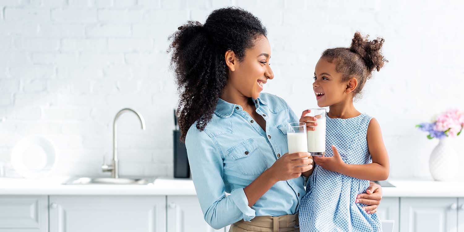 smiling african american mother and daughter holding glasses of milk in kitchen and looking at each other