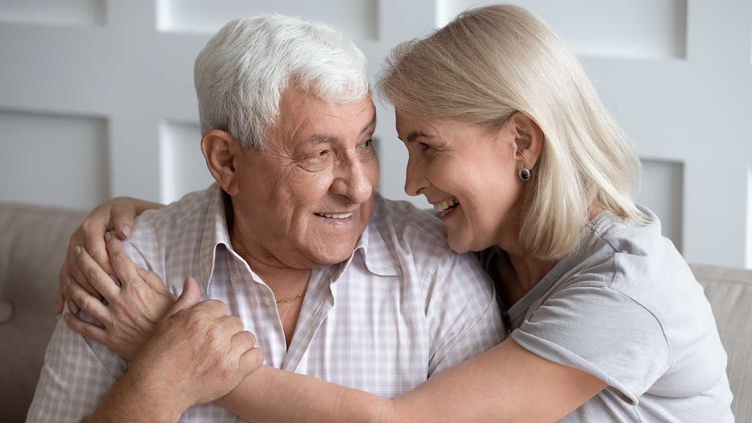 Happy older wife and husband hugging, looking in eyes, expressing love and support, family enjoying tender moment together, sitting on cozy sofa, mature father and middle-aged daughter close up