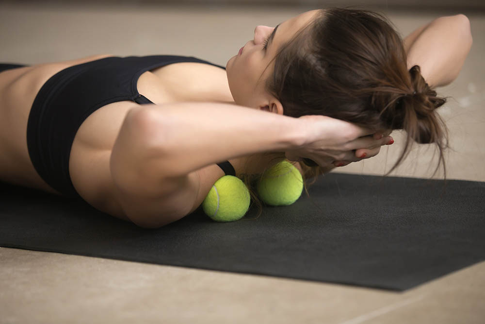 Lifestyle shot of young attractive female practicing self-massage technique applying tennis balls for neck and shoulder pain relief, working out on fitness mat on grey studio floor background, closeup