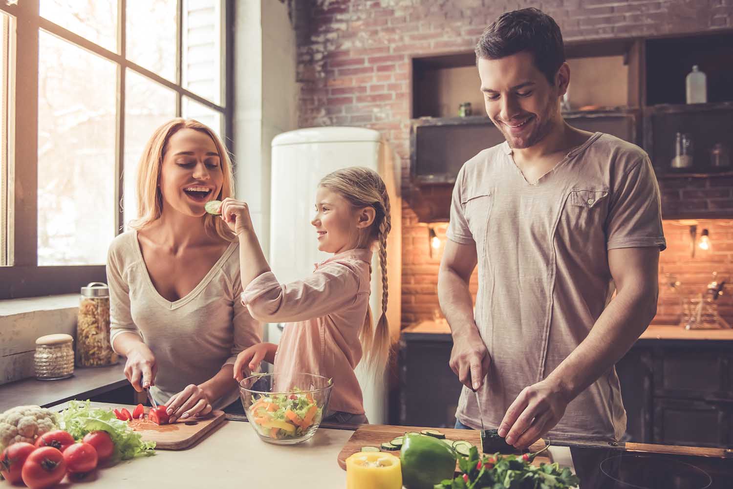 Cute little girl and her beautiful parents are cutting vegetables and smiling while cooking in kitchen at home
