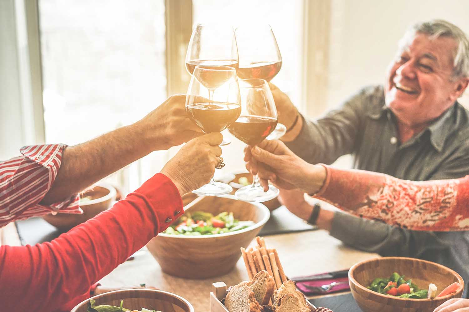 Senior friends cheering with wine glasses at home lunch - Happy mature people having fun together - Focus on left bottom glass - Joyful elderly lifestyle concept