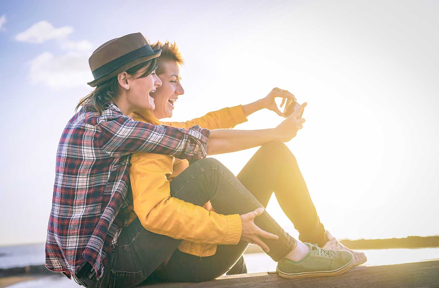 Happy lesbian couple taking a selfie with mobile smart phone camera on the beach at sunset - Vignette edit - Homosexuality, diversity, vacation, travel, lgbt, technology concept