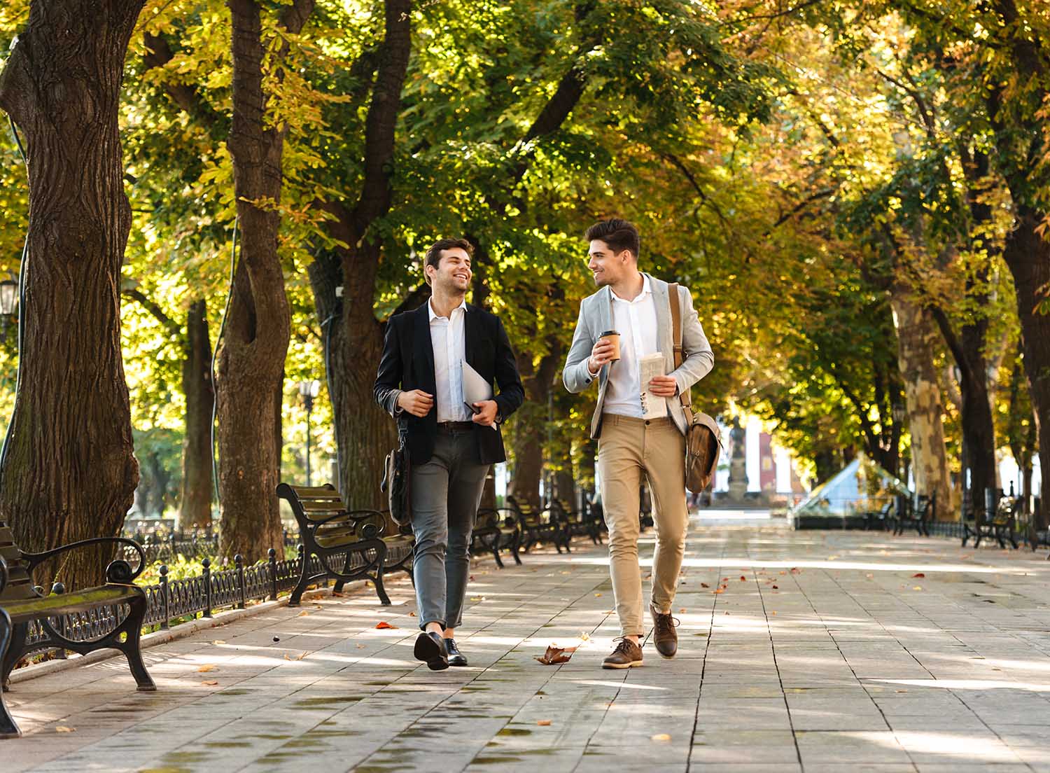Photo of young businessmen in suits walking outdoor through green park with takeaway coffee and laptop during sunny day