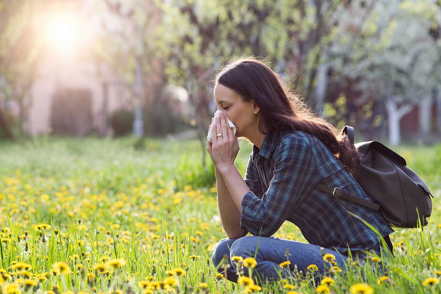 Young pretty woman blowing nose in grassland with spring flowers. Pollen allergy symptoms