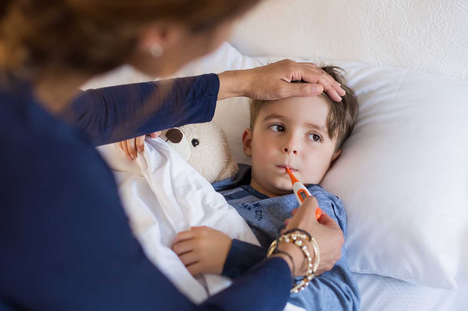 Sick boy with thermometer laying in bed and mother hand taking temperature. Mother checking temperature of her sick son who has thermometer in his mouth. Sick child with fever and illness in bed.