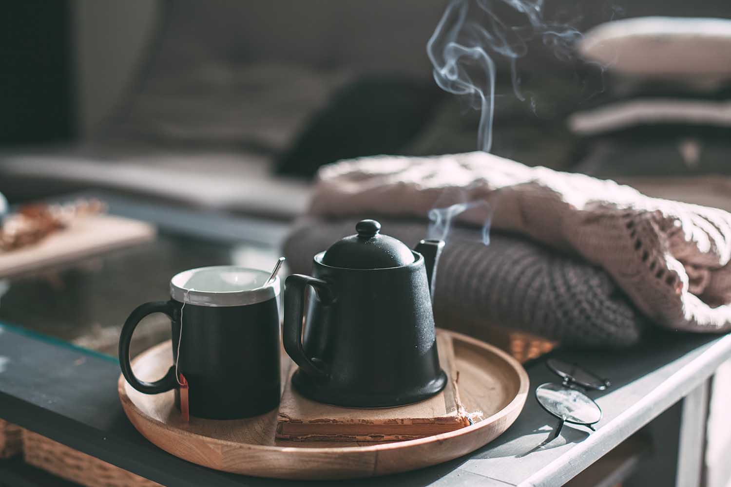 Still life details in home interior of living room. Sweaters and cup of tea with steam on a serving tray on a coffee table. Breakfast over sofa in morning sunlight. Cozy autumn or winter concept.