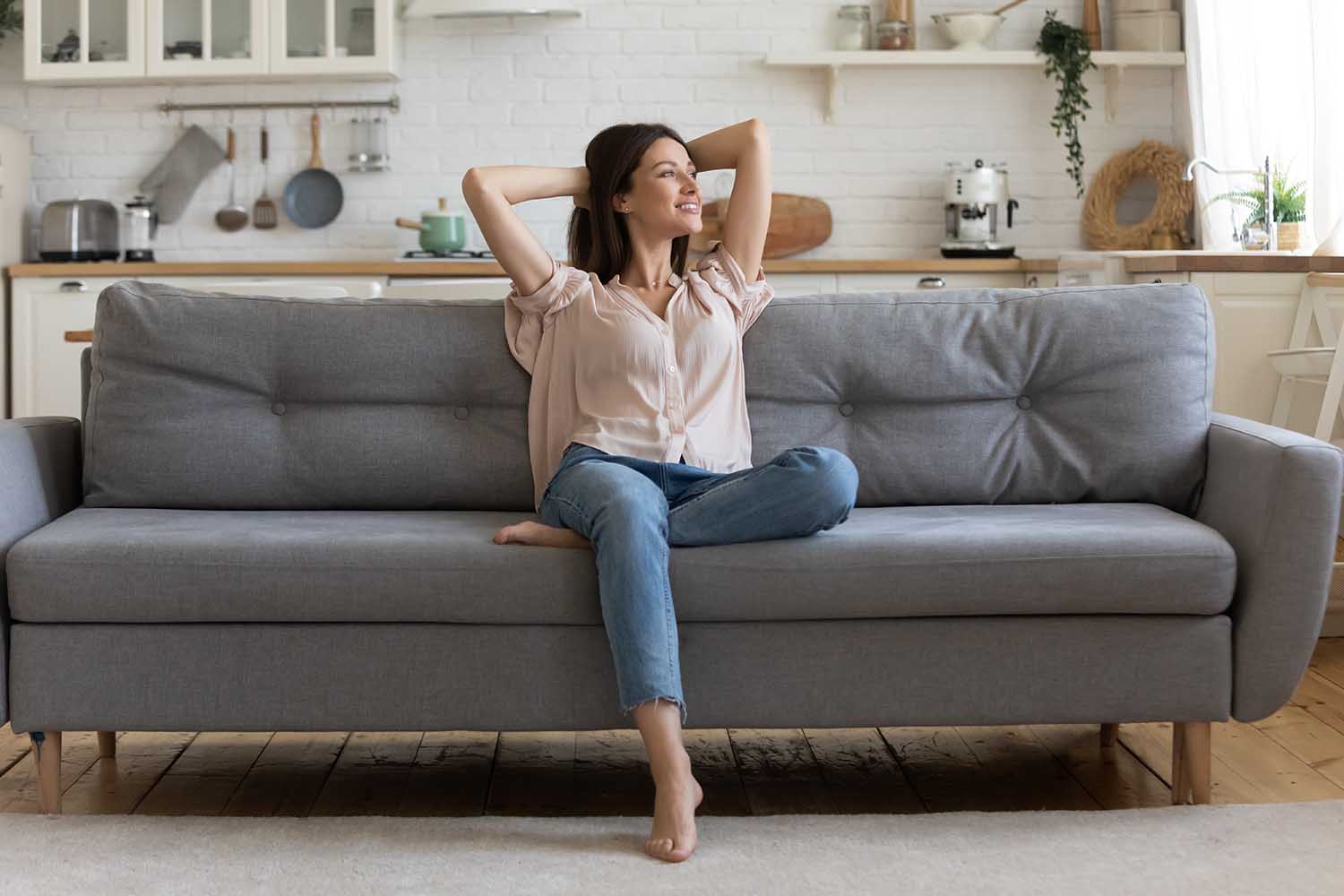 In cozy living room happy woman put hands behind head sitting leaned on couch 30s european female enjoy lazy weekend or vacation, housewife relaxing feels satisfied accomplish chores housework concept