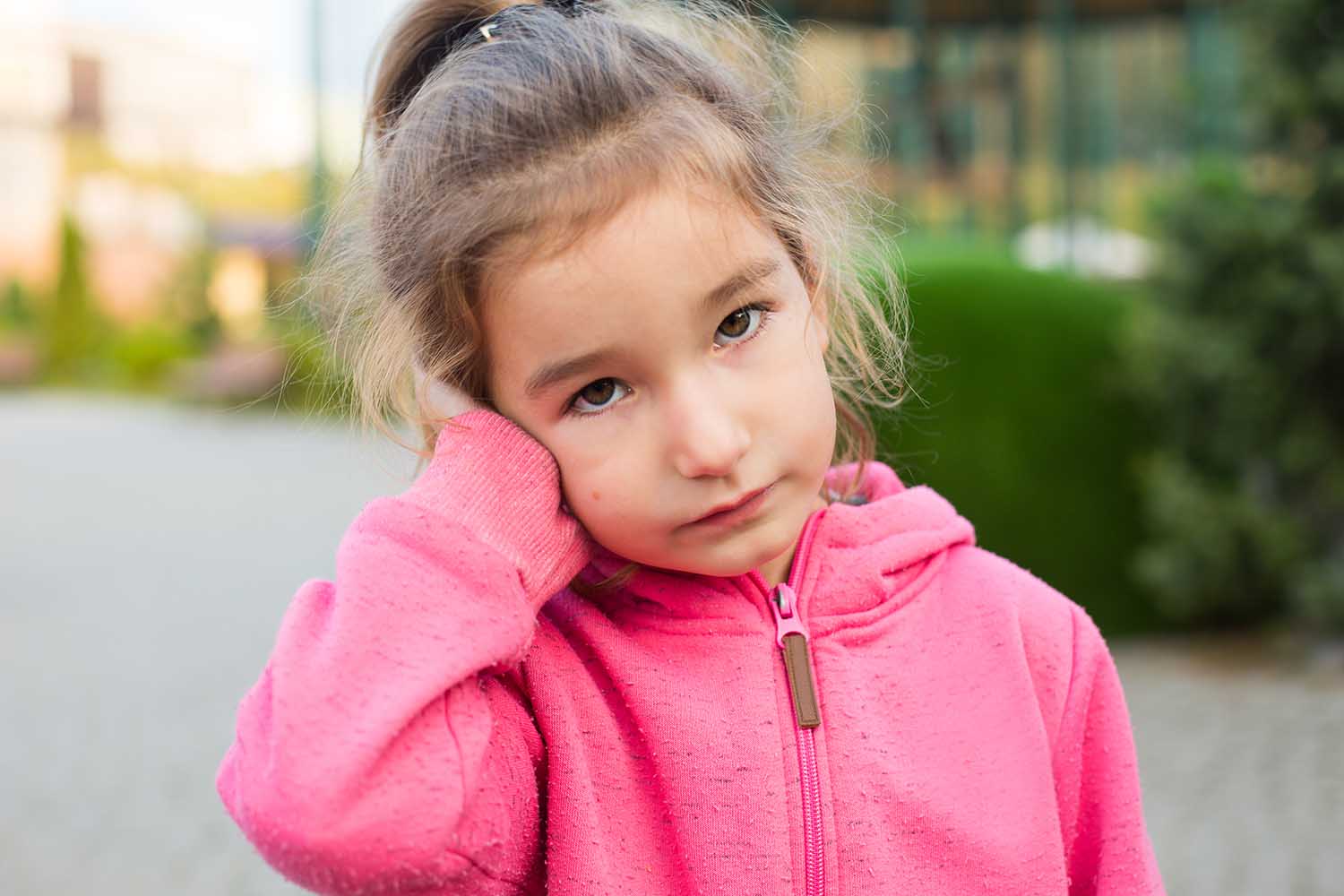 A little girl in a pink hoodie with a sad and tearful face is holding her ear. Ear pain, otitis media, swelling of the cheek, gums, toothache, children's surgery, otolaryngology. Children's medicine