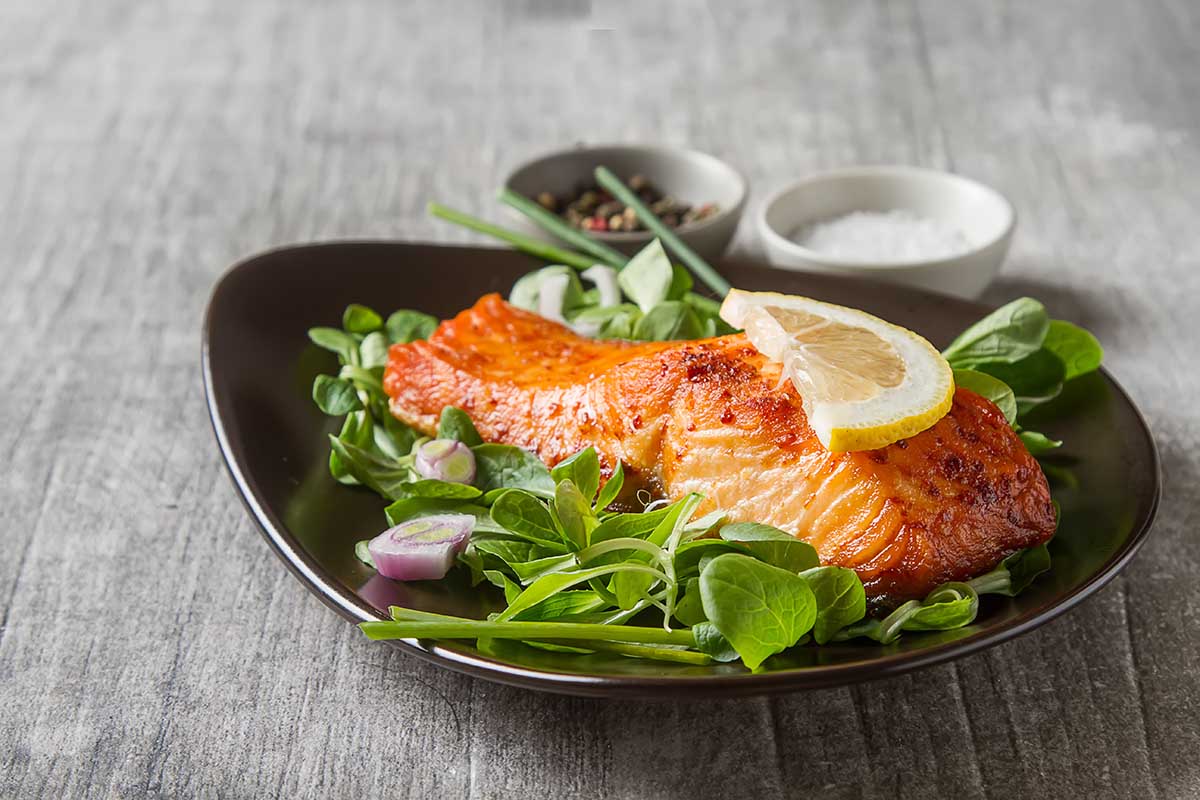 One piece of baked salmon grilled pepper lemon and salt on a brown plate with lettuce leaves. wood background