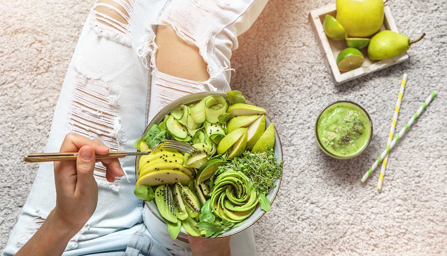 Woman in jeans holding fresh healthy greeen salad with avocado, kiwi, apple, cucumber, pear, greens and sesame on light background. Healthy food, clean eating, Buddha bowl salad, top view, detox
