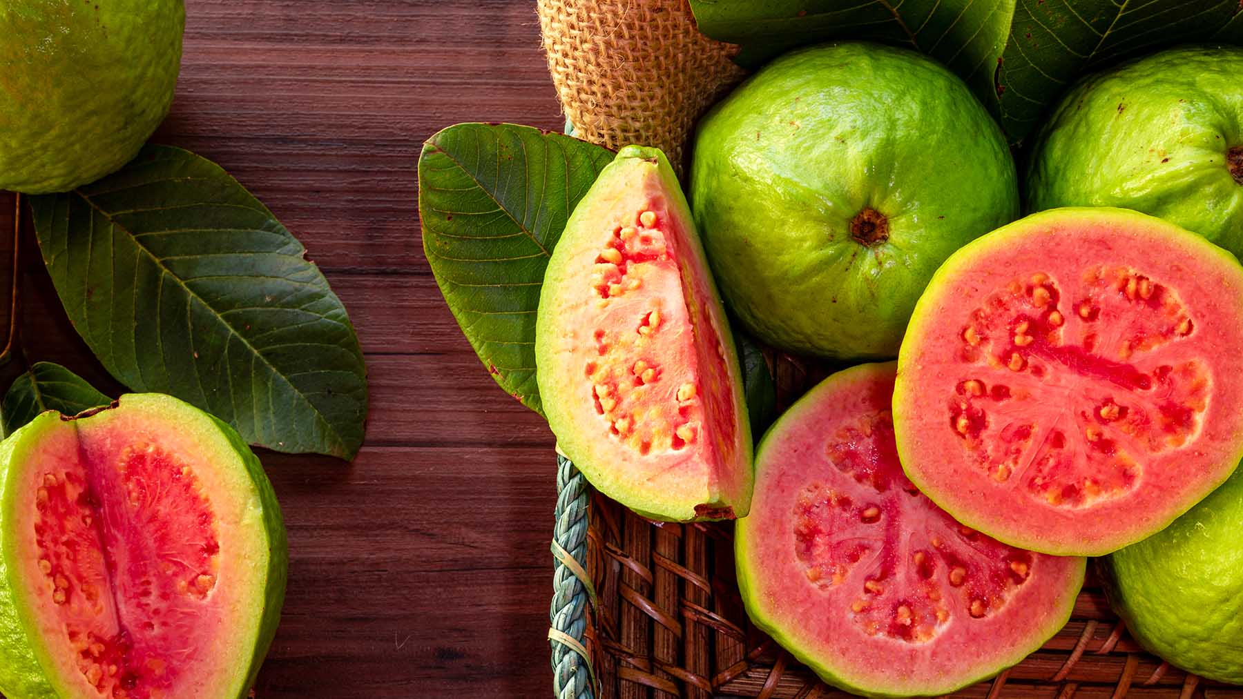Closeup of a red guava cut in half, in the background several guavas and green leaf