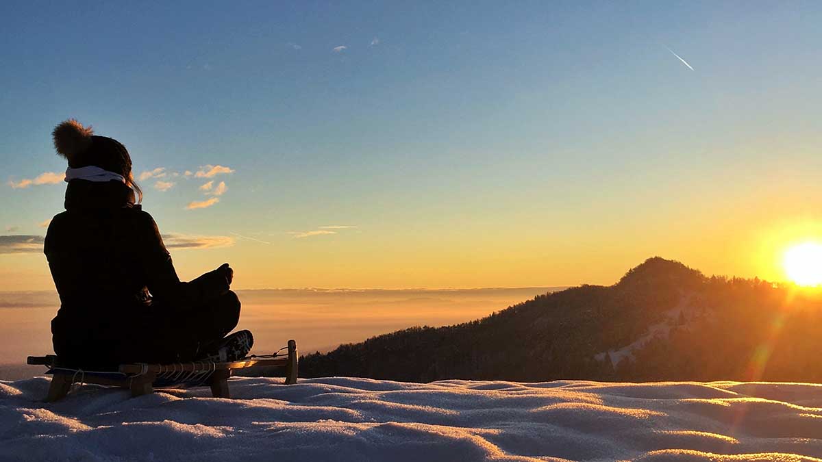 Young woman sitting and meditating on sledge looking at sunrise in winter.