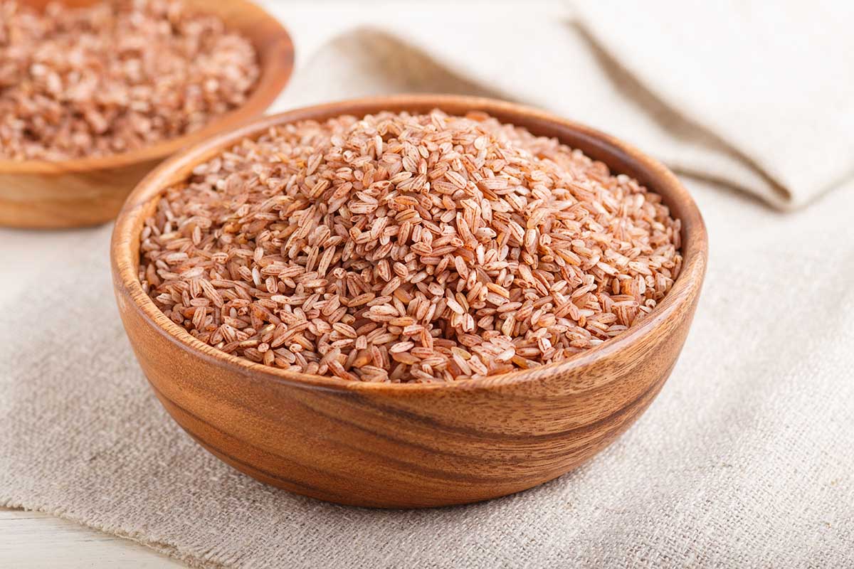 Two wooden bowls with unpolished brown rice on a white wooden background and linen textile. Side view, close up, selective focus.