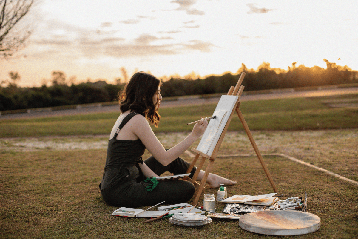 girl painting in a grass field with a sunset